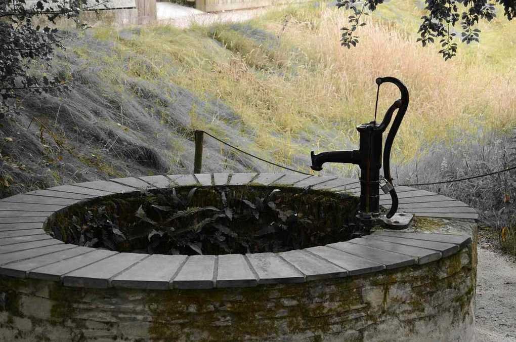 Un puits en pierre avec pompe manuelle, eau et colline herbeuse